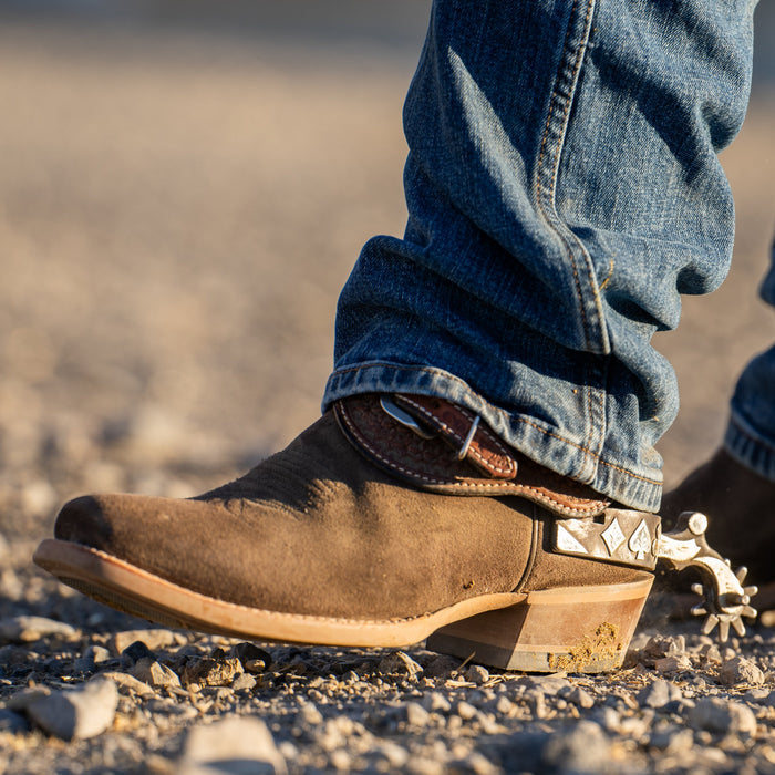 Brown cowboy boot with spurs on a gravel surface, wearing blue jeans.