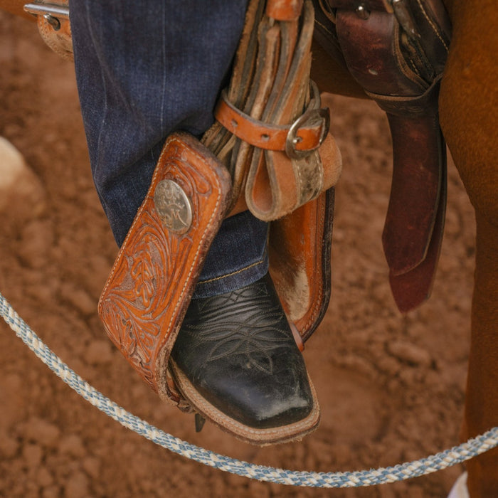Lifestyle image of black cowboy boot in a stirrup on a dirt background