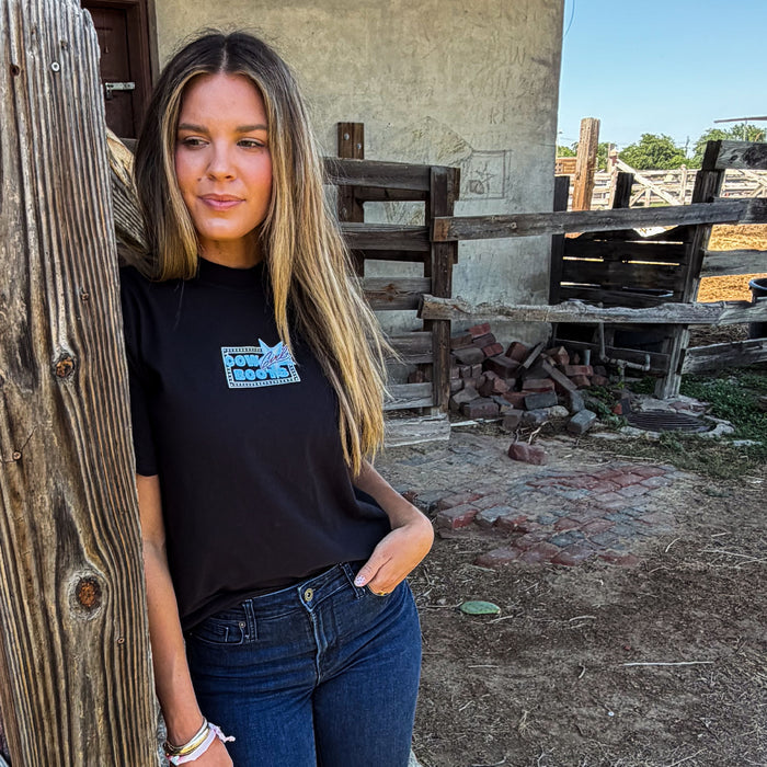 Woman wearing a black t-shirt with a logo, standing in front of rustic wooden structures.