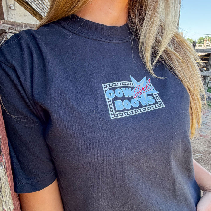 Black t-shirt with 'Cowgirl Boots' graphic worn by a person outdoors.