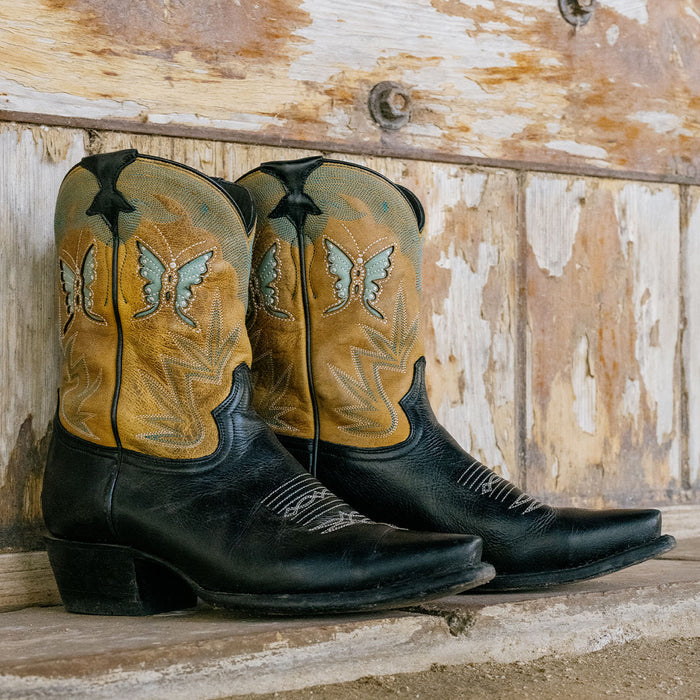 Lifestyle shot of black and tan cowgirl boots with butterfly inlays sitting against a distressed wooden wall background