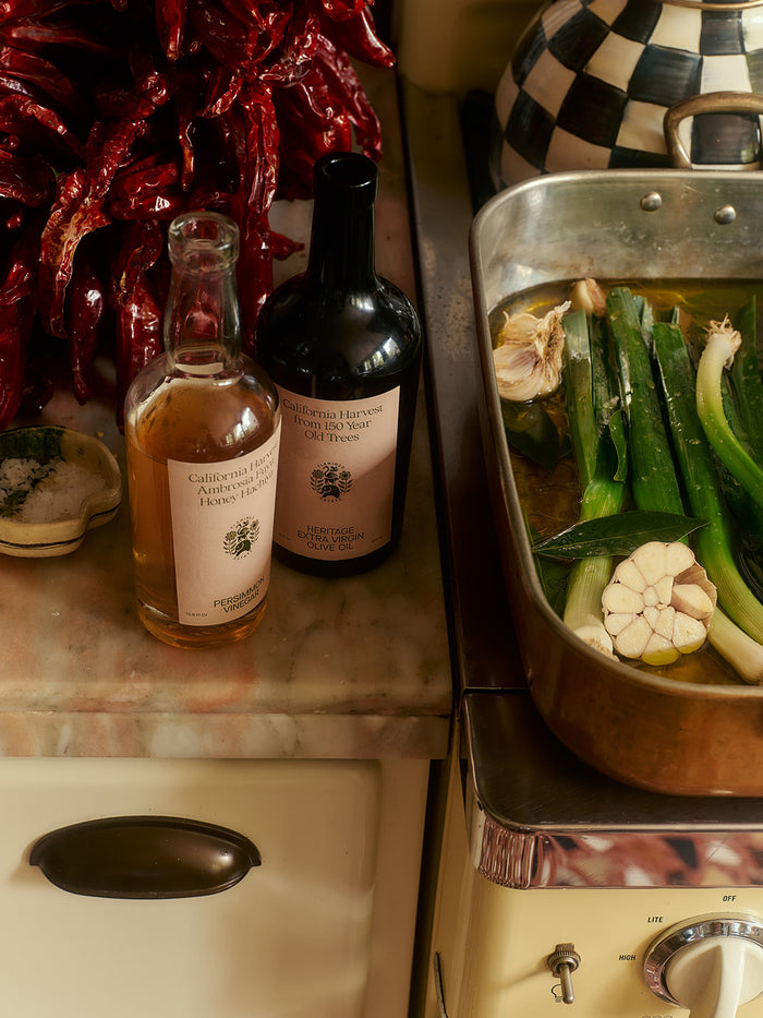 Bottles of Flamingo Estate vinegar and olive oil next to a roasting pan with garlic and leeks, set on a vintage kitchen countertop with dried red peppers in the background.