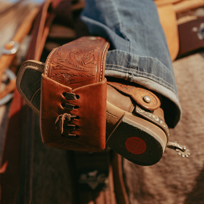 Close-up of a person wearing brown leather cowboy boots with intricate designs in a spur with horse blurred in background
